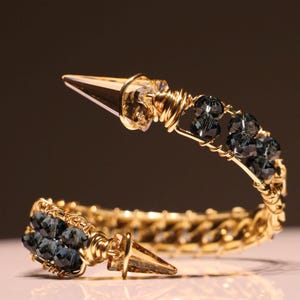 May include: A gold-toned bracelet with a spiked end and dark blue crystal beads. The bracelet features a woven design and is accented with gold wire wrapping. The spiked ends are a light brown color.