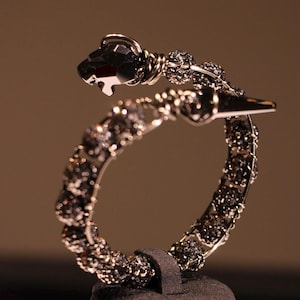 May include: A silver-toned bracelet with a black panther head design. The bracelet is adorned with black crystals and features a coiled design. The panther's head has a faceted, dark crystal eye. The bracelet is displayed on a black stand.