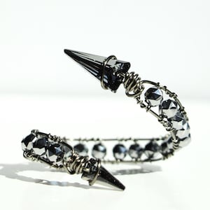 May include: A silver-toned cuff bracelet with a spiral design. The bracelet features faceted, gray crystal beads and two pointed, cone-shaped accents. The metal has a gunmetal finish, and the bracelet is set against a white background.