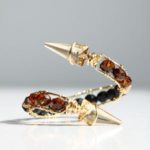 May include: A gold-toned bracelet with a spiral design. The bracelet features brown and black beads, and two gold-colored, pointed cone-shaped accents. The wire is intricately wrapped around the beads and cone-shaped accents.