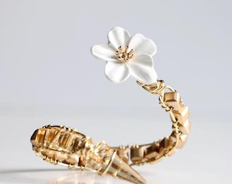 May include: A gold-toned bracelet with a white flower accent. The bracelet features a geometric design with a pointed end. The flower has a gold center. The bracelet is set against a white background.