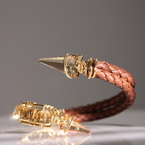 May include: A brown leather bracelet with gold-coloured metal accents. The bracelet has a braided leather band and is decorated with gold-toned spikes and crystal-like embellishments, creating an elegant design.