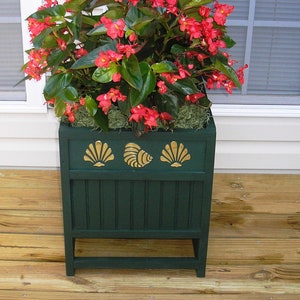May include: A green planter box with three gold seashell designs on the front. The planter is filled with a red flowering plant and is sitting on a wooden deck.