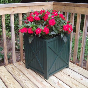 May include: A green wooden planter box with a criss-cross design on the front, filled with a vibrant red flowering plant. The planter is sitting on a wooden deck.