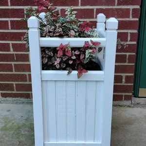 May include: A white wooden planter box filled with colorful plants. The box has a rectangular shape with vertical paneling on the front and sides. The plants have red, pink, and green leaves, adding a vibrant touch to the outdoor decor.
