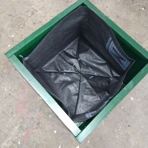 May include: A green wooden planter box with a black fabric liner inside. The liner is folded into a square shape.