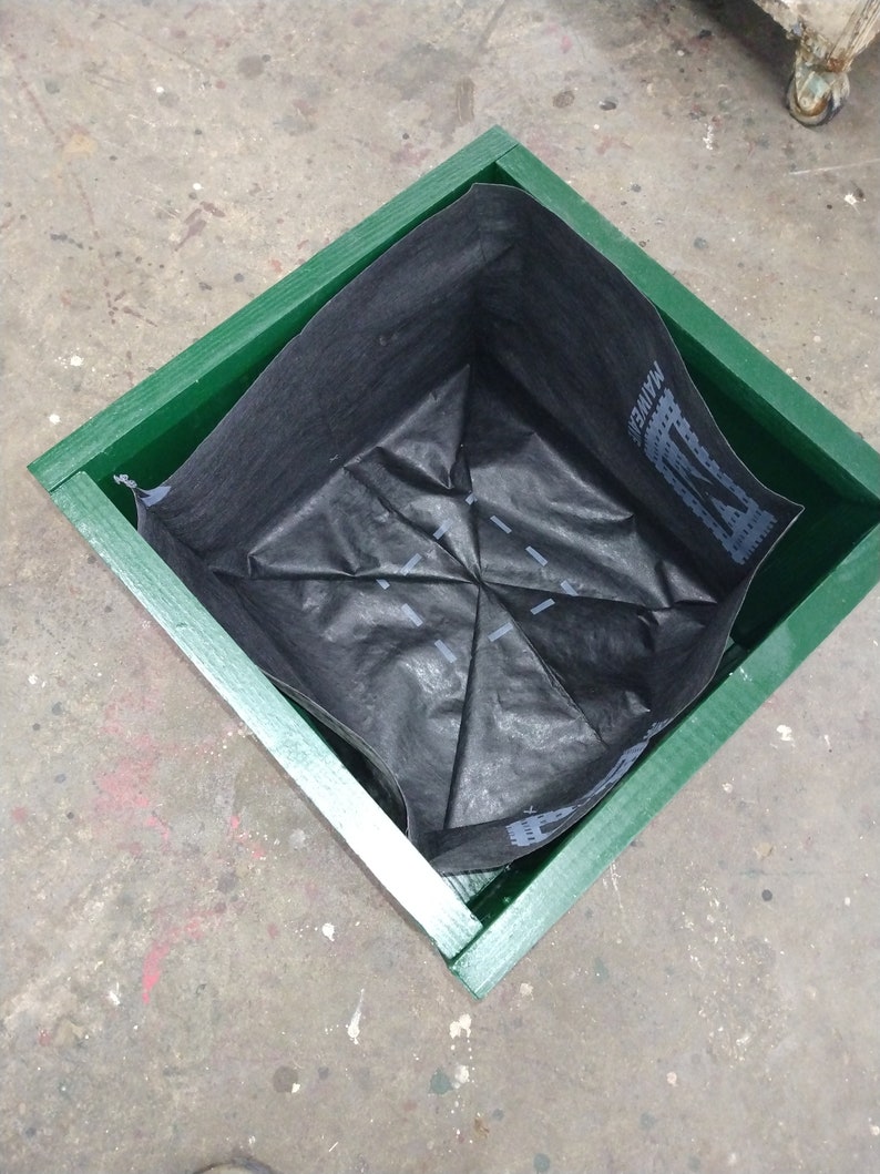 May include: A green wooden planter box with a black fabric liner inside. The liner is folded into a square shape.