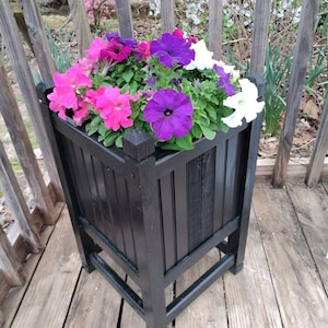 May include: A black wooden planter box with a square base and four legs, filled with a variety of colorful petunias, including pink, purple, and white.