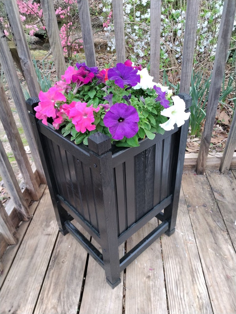 May include: A black wooden planter box with a square base and four legs, filled with a variety of colorful petunias, including pink, purple, and white.
