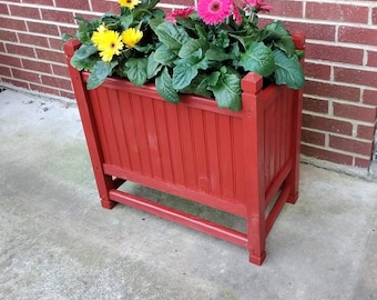 May include: A red wooden planter box with a decorative slatted design. The planter is filled with green plants and flowers, including pink and yellow gerbera daisies.