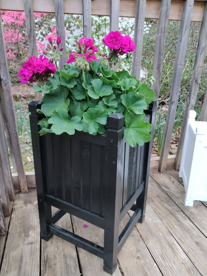 May include: A black wooden planter box with a square shape and four legs. The planter is filled with green plants and pink flowers.
