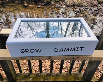 May include: A white rectangular planter box with a glass top, resting on a wooden structure. The words "GROW DAMMIT" are printed in black on the front of the box. The glass reflects the sky and trees.