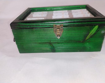 May include: A rectangular wooden storage box with a vibrant green stained finish and visible wood grain. The box features a hinged lid with a clear glass top and a central brass-coloured metal latch closure. The interior is divided into multiple compartments.