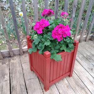 May include: A red wooden planter box with a square shape and vertical slats. The planter is filled with a green plant with bright pink flowers.
