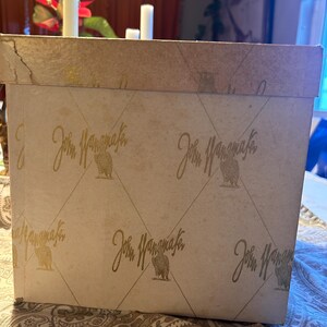 May include: A vintage rectangular box with a patterned design. The box is beige with a repeating gold pattern of a bird and the name "John Wanamaker" in a cursive font. The box has a slightly distressed appearance.
