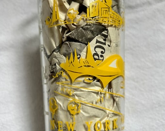 Puede incluir: Un vaso de vidrio transparente con un diseño amarillo que presenta una imagen estilizada del horizonte de la ciudad de Nueva York y el texto "NEW YORK WORLD'S FAIR 64". El vaso está lleno de papel de periódico arrugado.