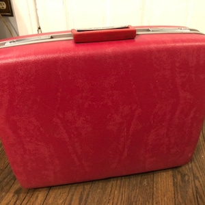 Könnte beinhalten: Ein vintage roter Hartschalenkoffer mit silbernen Akzenten und einem Griff.