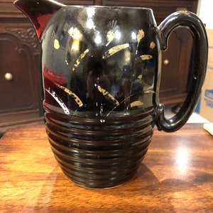 Puede incluir: Una jarra de cerámica negra con un diseño dorado. La jarra tiene una boca ancha y un asa. Tiene un diseño texturizado y acanalado en la parte inferior.