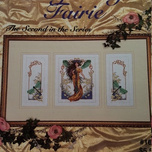 May include: A cross-stitch pattern book cover featuring a woman with long brown hair wearing a yellow dress and dragonfly wings. The title of the book is "DragonFly Fairie" and the subtitle is "The Second in the Series".