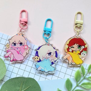 Puede incluir: Tres llaveros acrílicos con personajes de dibujos animados con vestidos rosas, azules y amarillos. Cada llavero tiene un cierre de diferente color: rosa, azul y beige. Los personajes tienen ojos grandes y están rodeados por un borde transparente.