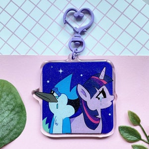 May include: A clear acrylic keychain featuring two cartoon characters against a starry blue background. The keychain has a purple heart-shaped clasp. One character is a blue bird, the other is a purple unicorn.