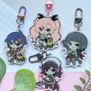 Op de afbeelding: Vier acryl sleutelhangers met anime-stijl karakters met metalen sluitingen. De karakters hebben verschillende haarkleuren en outfits, waarvan één een mes vasthoudt. De sleutelhangers staan op een roze en witte achtergrond met een groen blad.