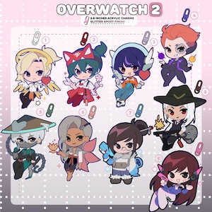 Op de afbeelding: Een set van negen Overwatch 2 acrylhangers, elk 6,35 cm groot met een glitter epoxy afwerking. De set bevat illustraties van personages in chibi-stijl, waaronder Mercy, Kiriko, Hana Song, Moira, Genji, Lifeweaver, Mei, Ashe en D.Va.