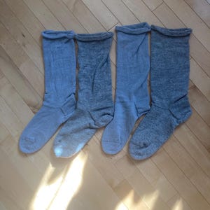 May include: Four pairs of gray crew socks are arranged on a light-colored wooden surface. The socks are made of a soft material and have a ribbed cuff at the top. The socks are in varying shades of gray.
