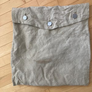 May include: A beige linen bag with a flap secured by three silver-colored buttons. The bag has a slightly wrinkled texture and a stain near the bottom. The bag is lying on a wooden surface.