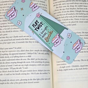 May include: A rectangular bookmark with a light green background and the text "PLOT TWIST: I bought MORE Books." The bookmark features illustrations of books and flowers. The bookmark is resting on an open book.