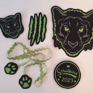 Könnte beinhalten: Schwarze und grüne Filzpatches mit einem Pantherkopf, Pfotenabdrücken, Krallen und einem grünen Band. Die Patches beinhalten auch einen schwarzen Kreis mit dem Text "Schulkind 2023" in grün.