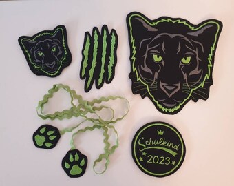 Könnte beinhalten: Schwarze und grüne Filzpatches mit einem Pantherkopf, Pfotenabdrücken, Krallen und einem grünen Band. Die Patches beinhalten auch einen schwarzen Kreis mit dem Text "Schulkind 2023" in grün.