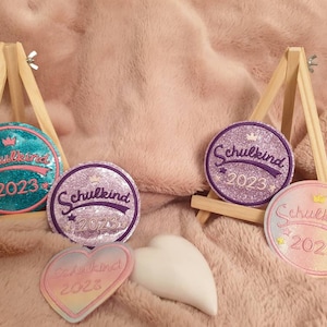 Könnte beinhalten: Drei glitzernde, runde Bügelbilder mit dem Text "Schulkind 2023" in einer bunten, Retro-Schrift. Die Patches sind in Türkis, Lila und Rosa mit Glitzerakzenten.