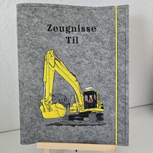 Könnte beinhalten: Grauer Filzordner mit einem aufgestickten gelben Bagger auf der Vorderseite. Der Ordner hat einen gelben Rand und der Text "Zeugnisse Til" ist auf der Vorderseite gestickt.