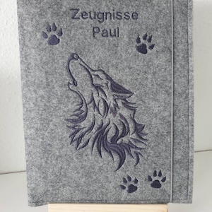 Könnte beinhalten: Ein grauer Filz-Dokumentenhalter mit dem Text "Zeugnisse Paul" in Dunkelblau gestickt. Der Einband zeigt einen heulenden Wolf und Pfotenabdrücke. Der Halter wird auf einer kleinen Holzstaffelei präsentiert.