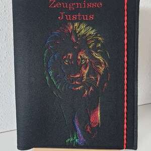 Könnte beinhalten: Schwarze Filzmappe mit einer roten Kordel am rechten Rand. Die Mappe hat einen bunten Löwen, der auf der Vorderseite gestickt ist, mit dem Text "Zeugnisse Justus" in Rot.