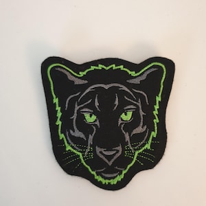 Könnte beinhalten: Schwarzer Panther-Aufnäher mit grünem Umriss und grünen Augen. Das Gesicht des Panthers ist auf einem schwarzen Aufnäher mit grünem Rand zentriert. Die Merkmale des Panthers sind mit grauen Nähten detailliert.