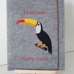 Könnte beinhalten: Grauer Filzordner mit einem roten und grünen Band. Der Ordner hat einen gestickten Tukan mit gelbem Schnabel und die Worte "Zeugnisse" und "Malte Jonah" in rot.