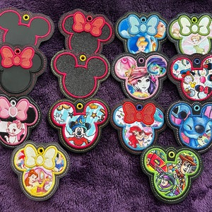 May include: A collection of Mickey Mouse-shaped keychains with various designs. Some feature solid black or glittery black faces with bows, while others showcase Disney characters like Cinderella, Ariel, and Stitch. Each keychain has a black border and a yellow loop.