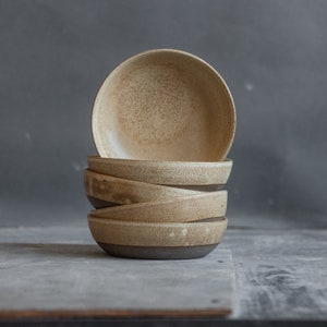 Puede incluir: Una pila de cinco cuencos de cerámica hechos a mano. Los cuencos tienen un interior beige claro moteado y un exterior marrón mate más oscuro. Los cuencos están apilados uno encima del otro, mostrando su diseño simple pero elegante. Perfectos para servir sopas, ensaladas o aperitivos.