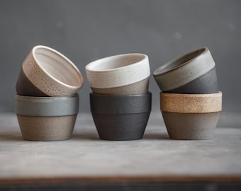 Peut inclure: Quatre tasses en céramique empilées en trois combinaisons de couleurs différentes. Les tasses sont dans des tons de gris, blanc et marron. Les tasses sont disposées en rangée sur une surface grise.