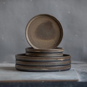 Może przedstawiać: Stos sześciu brązowych ceramicznych talerzy z ciemną obwódką. Talerze są okrągłe z lekko podniesioną krawędzią. Talerze są ułożone jeden na drugim.