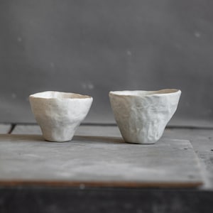 May include: Two handmade, white ceramic cups with textured surfaces. The cups have an organic shape and a speckled appearance. The cups are displayed on a grey surface, with a dark grey background.