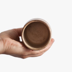 Op de afbeelding: Een beige keramische beker met een gespikkeld glazuur. De beker is leeg en wordt vastgehouden door een hand.