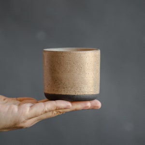 Op de afbeelding: Een kleine, beige keramische mok met een donkerbruine rand. De mok heeft een gespikkeld, getextureerd oppervlak.