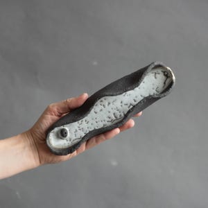 Może przedstawiać: Wydłużona, ręcznie formowana taca ceramiczna o szorstkiej, ciemnoszarej powierzchni zewnętrznej i kontrastującym, gładkim białym wnętrzu z drobnymi ciemnymi plamkami oraz pojedynczym okrągłym wgłębieniem na jednym z końców naczynia.