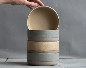 May include: A stack of three ceramic bowls with a cylindrical shape. The top bowl is beige with a speckled texture, the middle bowl is beige, and the bottom bowl is a muted sage green. Each bowl features a smooth, matte finish and straight sides.