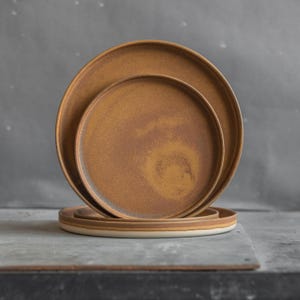 Puede incluir: Tres platos de cerámica de diferentes tamaños, apilados uno encima del otro. Los platos son de color marrón oxidado con un acabado moteado.