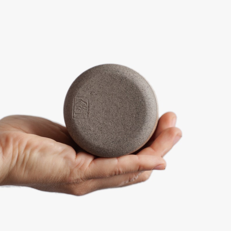Op de afbeelding: Een ronde, grijze keramische pot met een getextureerd oppervlak. De pot heeft een klein, rechthoekig merkteken op de bodem met de tekst "KIMI".