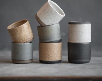 Peut inclure: Quatre tasses en céramique empilées en deux paires. Les tasses sont toutes de couleurs différentes et ont un glaçage moucheté. Les tasses sont sur une surface grise.